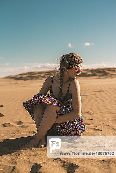 Frau mit Sonnenbrille sitzt am Strand im Sand vor blauem Himmel am sonnigen Tag