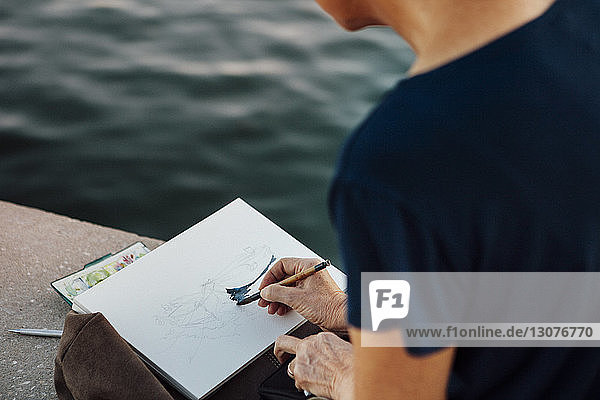 Beschnittenes Bild einer Frau  die auf Papier malt  während sie an einer Stützmauer sitzt