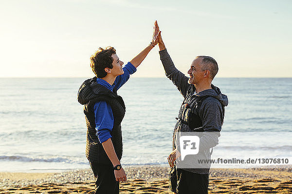 Vater und Sohn machen High-Five  während sie bei Sonnenuntergang am Strand gegen Meer und Himmel stehen
