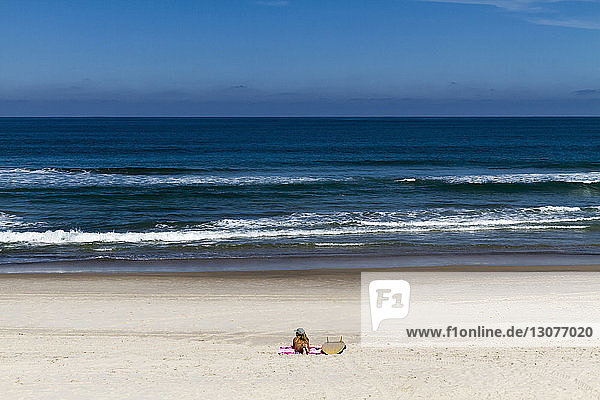 Mitteldistanzansicht einer Frau  die am Strand am Surfbrett auf einer Decke vor blauem Himmel am sonnigen Tag liegt