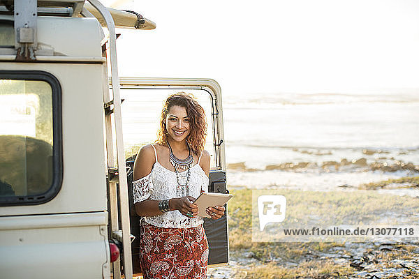 Portrait of happy woman with tablet computer standing by off-road vehicle at beach