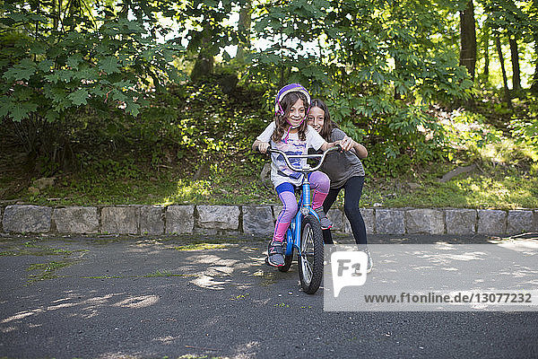 Girl assisting sister in riding bicycle on road against trees at park