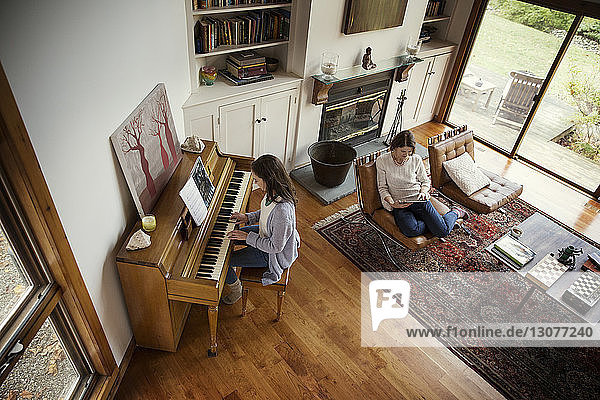 Hochwinkelansicht einer Tochter  die Klavier spielt  und einer Mutter  die einen Tablet-Computer im Wohnzimmer benutzt