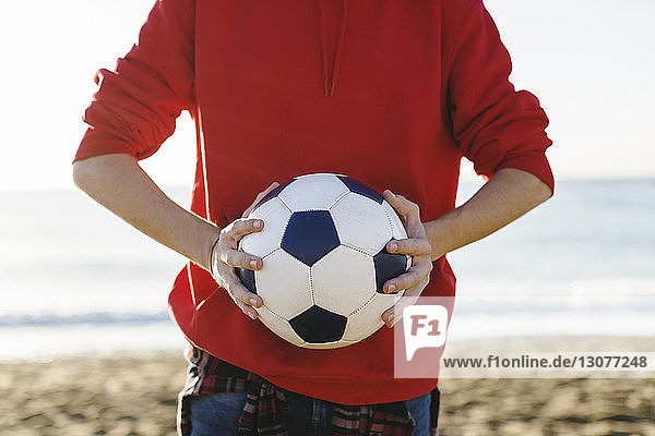 Midsection of teenage boy holding soccer ball while standing at beach against sea