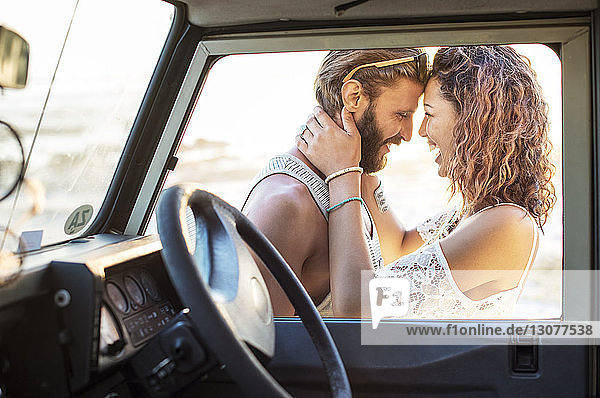 Cheerful couple looking at each other while standing by off-road vehicle seen through window