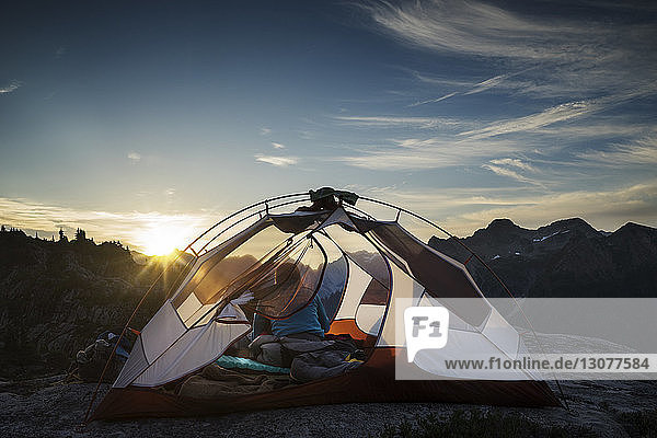 Rückansicht einer Frau  die bei Sonnenuntergang im Zelt vor den Bergen sitzt