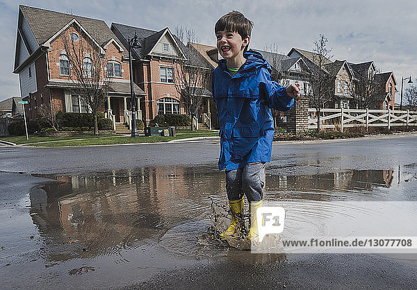 Full length of playful boy wearing rubber boots while jumping in puddle on road against houses