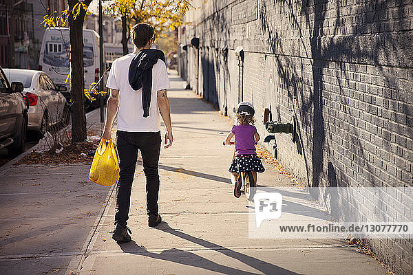Rear view of father walking while daughter cycling on footpath