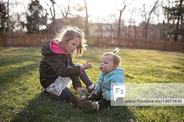 Sisters blowing bubbles while sitting on grassy field at park