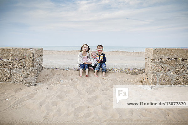 Portrait of siblings sitting at beach against cloudy sky