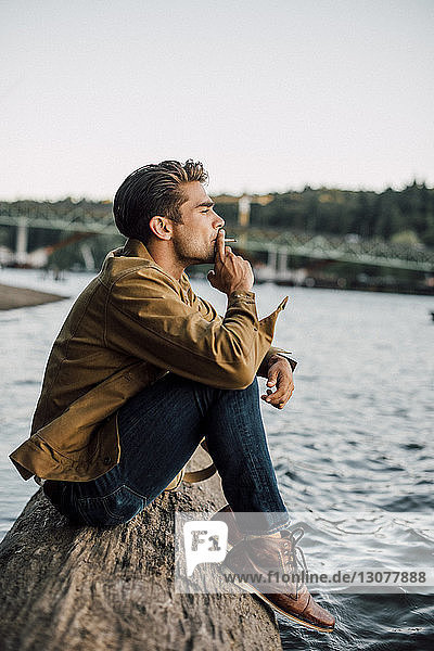 Side view of man smoking while sitting on log by river against clear sky