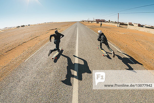 Full length rear view of friends skateboarding on road against sky during sunny day
