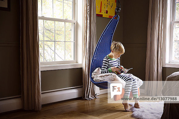 Junge liest in voller Länge ein Buch  während er auf einer Schaukel im Schlafzimmer sitzt