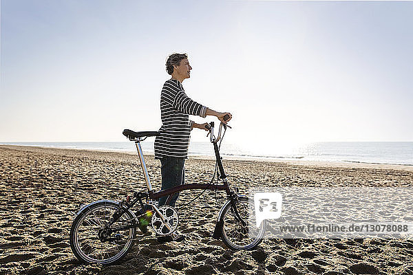 Frau steht mit Fahrrad am Strand vor klarem Himmel