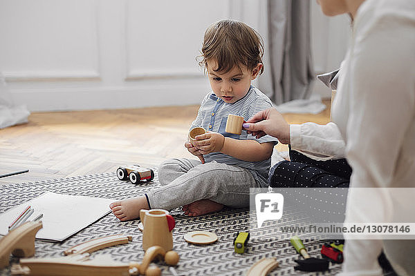 Mittendrin gibt die Mutter dem Jungen eine Spielzeug-Teetasse  während sie zu Hause auf dem Teppich sitzt