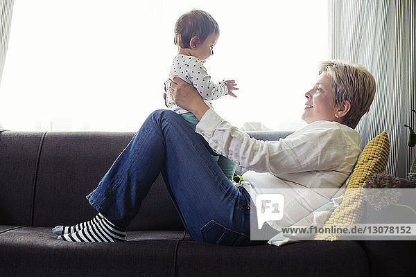 Seitenansicht einer lächelnden reifen Frau  die mit ihrer Enkelin auf dem Sofa am Fenster spielt