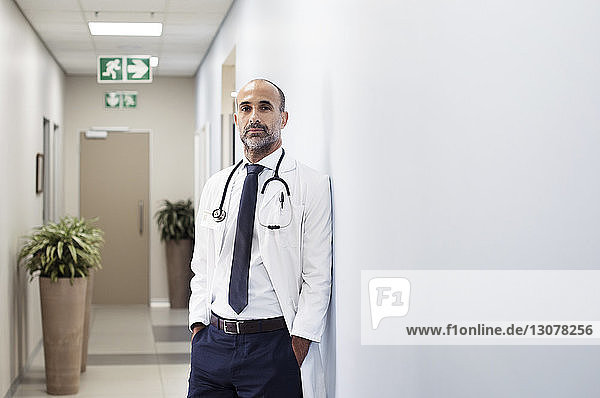 Porträt eines Arztes mit an die Wand gelehnten Händen in Taschen im Krankenhaus