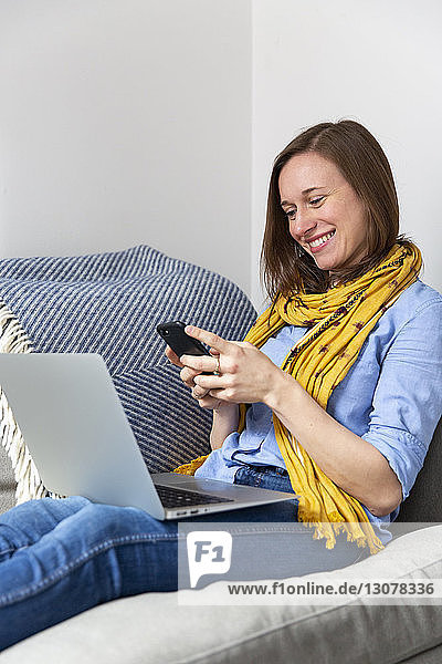 Lächelnde Frau benutzt Mobiltelefon  während sie mit Laptop-Computer auf dem Sofa im Wohnzimmer sitzt