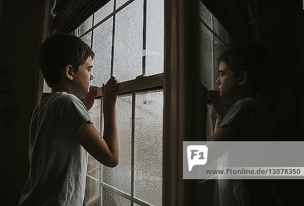 Seitenansicht eines nachdenklichen Jungen  der durch ein nasses Fenster schaut und sich zu Hause im Spiegel spiegelt