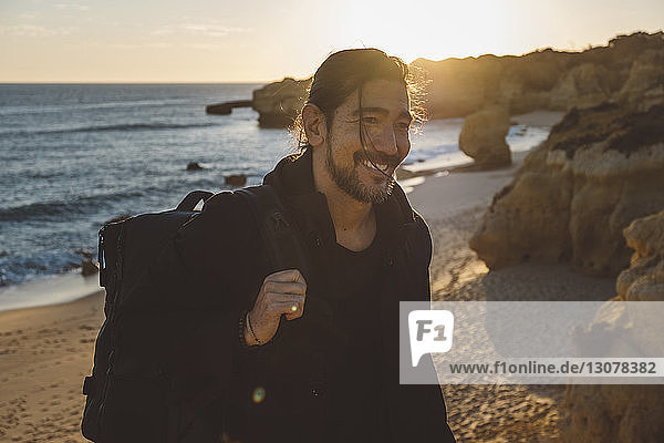 Smiling man with backpack standing at beach against sky during sunset
