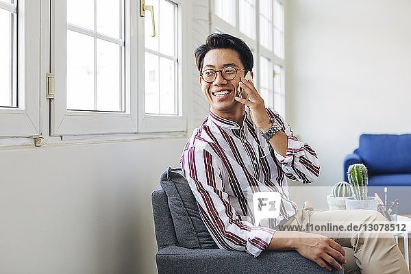 Happy businessman talking on smart phone while looking through window in office
