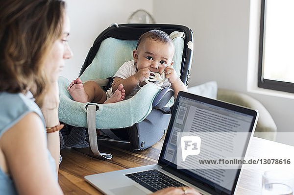 Mother using laptop computer by baby boy in seat on table at home