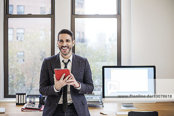 Happy businessman using tablet computer while leaning on desk in office