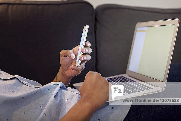 Seitenansicht eines Illustrators mit Smartphone und Laptop im Mittelschnitt  während er im Kreativbüro auf dem Sofa liegt