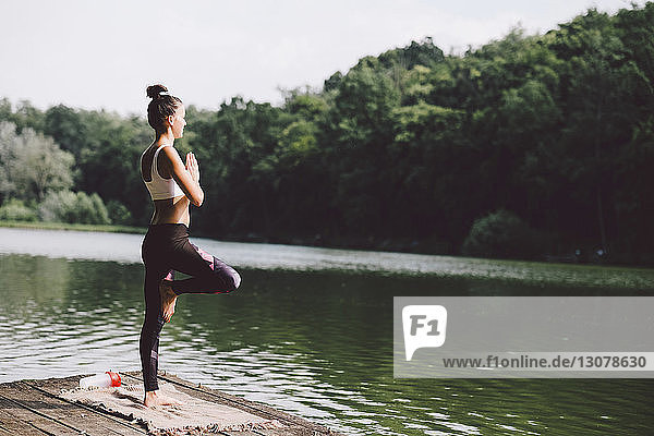 Seitenansicht einer Frau  die meditiert  während sie auf einem Steg über einem See im Wald steht