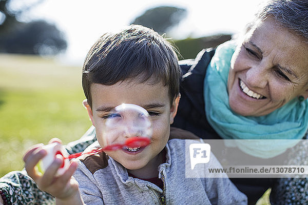 Glückliche ältere Frau schaut auf ihren Enkel  der im Park mit dem Blasenzauberstab spielt