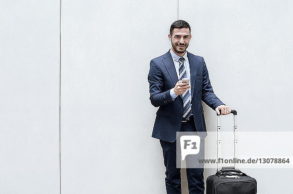 Portrait of happy businessman holding smart phone and luggage against wall