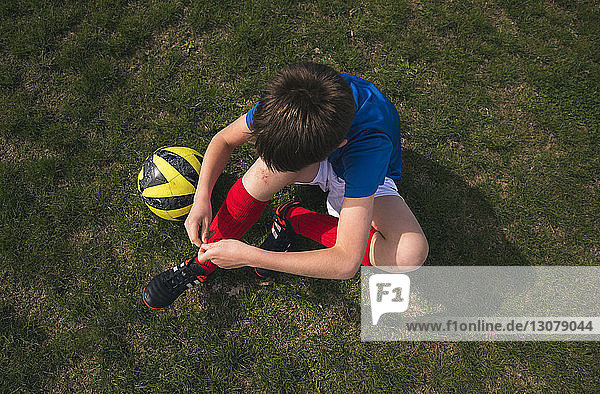Schrägansicht eines Jungen beim Schnürsenkelbinden  der auf einem Fussballfeld sitzt