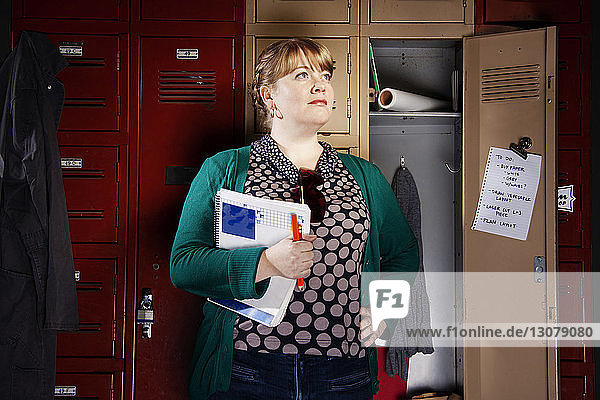 Confident woman holding papers looking away while standing against cabinet in office