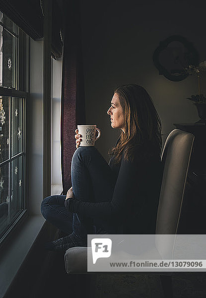 Nachdenkliche Frau mit Kaffeetasse  die durch das Fenster schaut  während sie zu Hause sitzt
