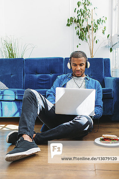 Mann in voller Länge  der Musik über einen Laptop-Computer hört  während er auf dem Boden im Wohnzimmer sitzt
