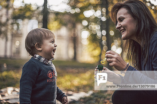 Mutter hält Seifenblasen-Stab  während sie den fröhlichen Sohn betrachtet  der im Herbst im Park spielt