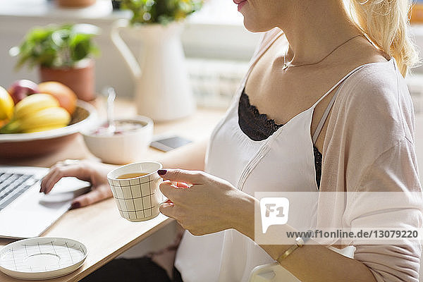 Mittelsektion einer Frau  die am Tisch sitzend mit einem Laptop Kaffee hält