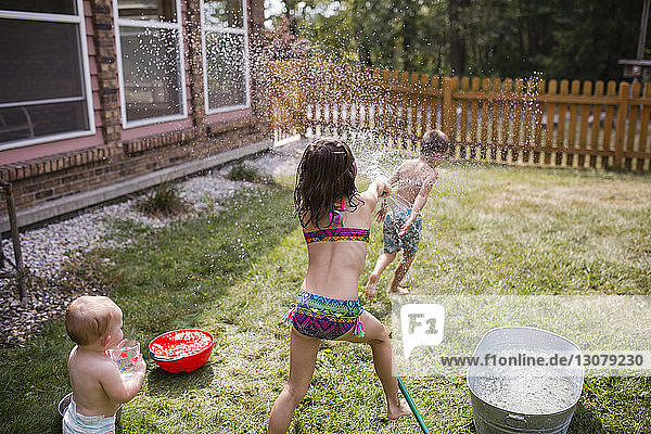 Baby girl looking at sister spraying water on shirtless brother through hose
