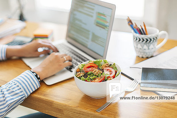 Beschnittenes Bild einer Geschäftsfrau  die am Laptop-Computer an einer Salatschüssel auf einem Holztisch im Heimbüro bloggt