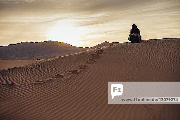 Rückansicht einer Frau  die auf Sand in der Wüste vor bewölktem Himmel bei Sonnenuntergang sitzt