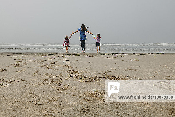 Rückansicht einer Mutter  die die Hände ihrer Töchter hält  während sie am Strand gegen den Himmel steht