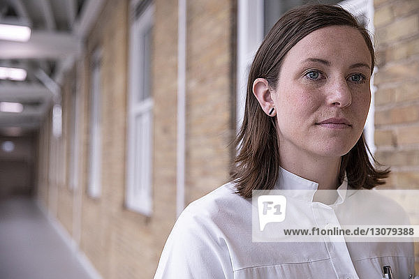 Close-up of thoughtful female doctor looking away while standing against brick wall in hospital corridor