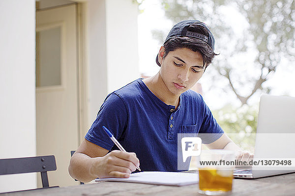 Junger Mann schreibt in Buch  während er mit seinem Laptop am Tisch im Hof sitzt