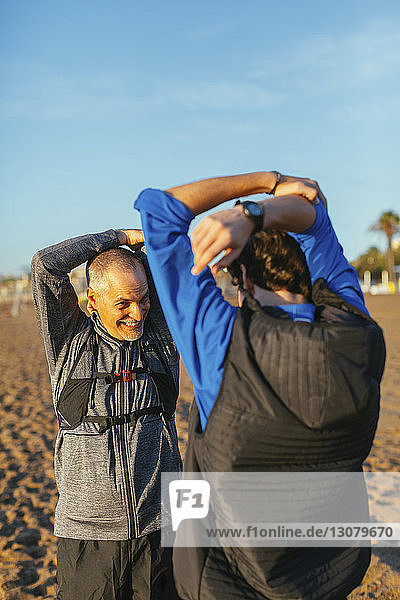 Father and son stretching arms while standing face to face at beach against sky during sunset