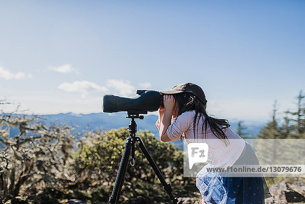 Side view of girl looking through telescope while standing against sky