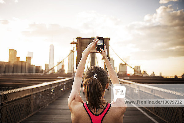 Rückansicht einer Frau  die die Brooklyn-Brücke per Mobiltelefon fotografiert