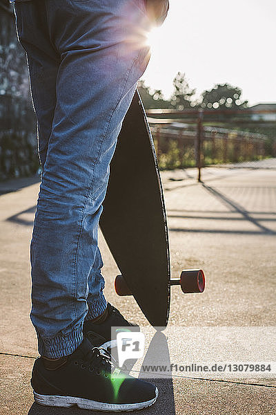 Niedriger Teil eines Mannes  der ein Skateboard hält  während er bei Sonnenuntergang gegen den Himmel steht