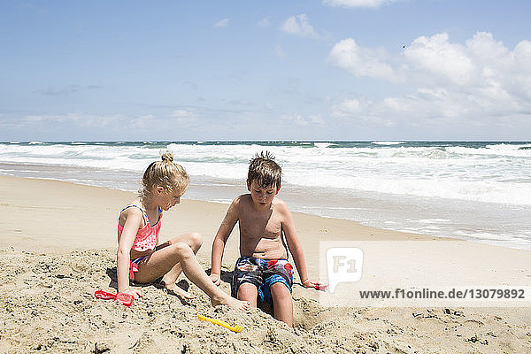 Geschwister spielen mit Sand am Strand gegen den Himmel während eines sonnigen Tages