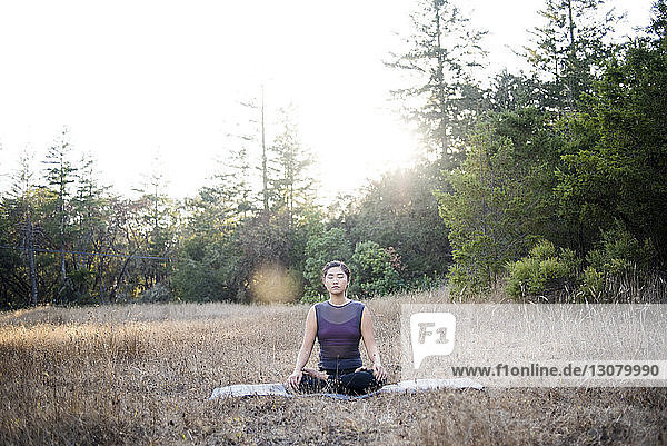 Frau meditiert  während sie auf einer Übungsmatte vor klarem Himmel auf dem Feld sitzt
