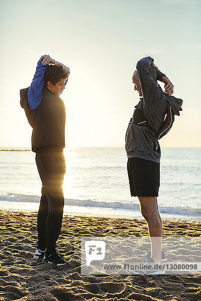 Full length of father and son stretching arms while standing face to face at beach against clear sky during sunset
